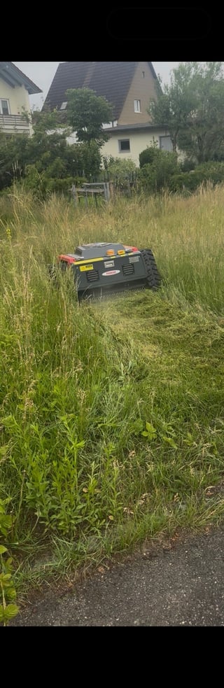 Robotic lawn mower with red and yellow accents cutting tall grass in residential yard
